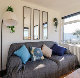 A living room with a sofa covered with a dark fabric and cushions by a window with a lamp and three vertical mirrors on the wall at Seacroft in Crantock