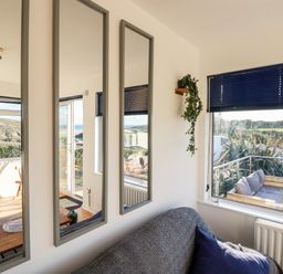 A room with a sofa covered by a throw and a blue pillow near a window with blue blinds and three vertical mirrors on the wall at Seacroft in Crantock