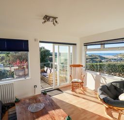 A sunlit living room with wooden rocking chair papasan chair wooden table and large windows overlooking outdoor seating and landscape at Seacroft in Crantock