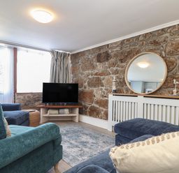 A living room with a stone wall a round mirror blue armchairs a television and striped curtains at Harbour Studio in Sennen Cove