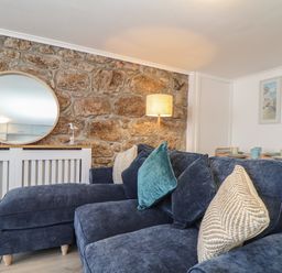 A living room with a blue sectional sofa stone wall a round mirror a floor lamp and a dining table at Harbour Studio in Sennen Cove