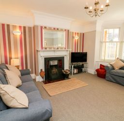 A living room with gray sofas a fireplace a wall mirror a television on a glass stand and a window with blinds at Westholme Lodge in Minehead