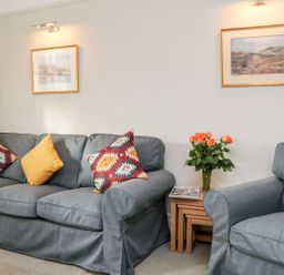 A living room with a sofa and armchair with cushions a side table with flowers and magazines and framed pictures on the wall at Westholme Lodge in Minehead
