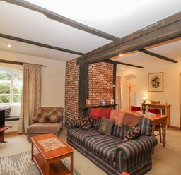 A living room with striped sofa, wooden coffee table, TV, armchair, dining table, exposed wooden beams, and brick column at Field Cottage in Shepton Mallet
