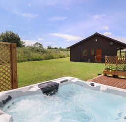 An outdoor hot tub on a wooden deck with a wooden cabin and green lawn in the background at Lodge 1 in South Hykeham