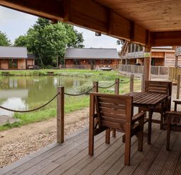 A covered wooden deck with table and chairs overlooking a pond and wooden lodges at Lodge 24 in South Hykeham