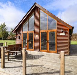 An outdoor wooden deck with a table and benches in front of a wooden lodge with large windows at Lodge 3 in Thorpe-On-The-Hill