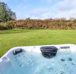 An outdoor hot tub filled with water on a grassy lawn with trees and bushes in the background at Lodge 3 in Thorpe-On-The-Hill