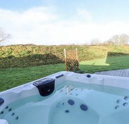 An outdoor hot tub filled with water on a wooden deck with grass and bushes in the background at Lodge 7 in South Hykeham