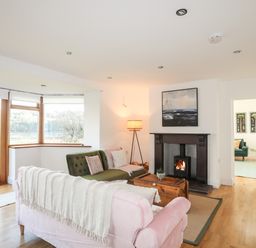 A living room with a pink sofa, green armchair, wooden chest coffee table, fireplace, and large windows at Ynys Faelog Cottage in Ynys Faelog near Menai Bridge
