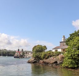 A house surrounded by trees on a rocky shoreline with boats and a suspension bridge in the background at Ynys Faelog near Menai Bridge