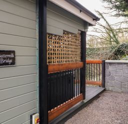 An outdoor area with a grey wooden wall and a sign reading Buttermilk Bridge next to a gravel path and wooden railing at Buttermilk Bridge in Dungiven