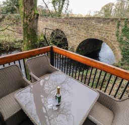 A balcony with a glass table and wicker chairs overlooking a stone bridge and river at Buttermilk Bridge in Dungiven
