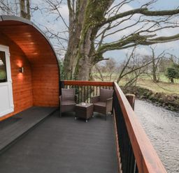 A wooden cabin balcony with two wicker chairs and a small table overlooking a river in a rural area at Pine in Dungiven Londonderry