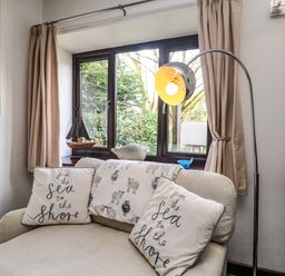 A beige armchair with two cushions and a sheep-patterned blanket near a window with beige curtains and a standing lamp at The Old Piggeries in Gulval near Penzance
