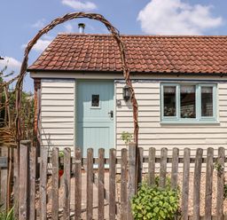 A small house with a light blue door and windows behind a wooden fence with a woven arch at The Calf Pen in Colyton