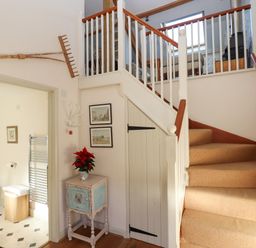 A staircase with beige carpet and wooden handrails next to a small blue cabinet with a red plant and two framed pictures on the wall at The Hay Loft in Colyton