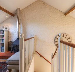 An attic room with a wood stove a beige ottoman a bookshelf a television and a large wall clock at The Hay Loft in Colyton