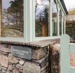 Stone wall with a sign reading Scandale Bridge Cottage next to a window with green frames and an outdoor patio with chairs at Scandale Bridge Cottage Ambleside