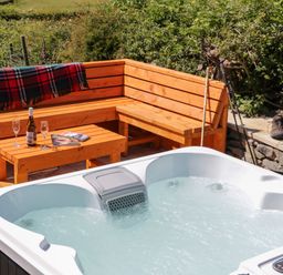 An outdoor seating area with wooden benches a table with a bottle and glasses and a hot tub at Holly in Ulverston