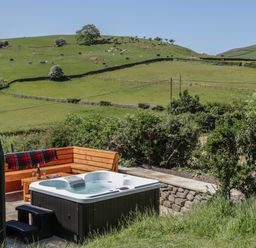An outdoor hot tub next to wooden seating and green fields with grazing cows at Rowan in Ulverston