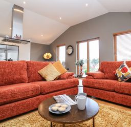 A living room with two red sofas a round coffee table with mugs and pastries a kitchen area and windows with blinds at Woodbridge Penthouse in Woodbridge