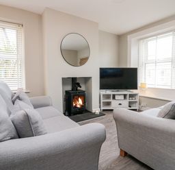 A living room with two grey sofas a wood-burning stove a round mirror a television on a white stand and windows with blinds at Wainwright Cottage in Gosforth