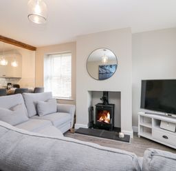 A living room with gray sofas a wood stove a round mirror a television and an open view into a kitchen at Wainwright Cottage in Gosforth