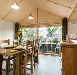 A dining area with wooden table and chairs set with plates and glasses beside a glass door with outdoor seating at Lodge 3 in Corfe Castle