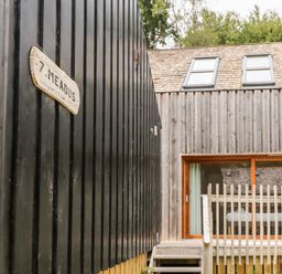 A wooden house with large windows and two roof skylights next to a black wooden wall with a sign reading 7 Meads at Lodge 6 in Corfe Castle