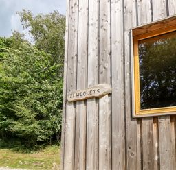 A wooden exterior wall with a window and a wooden sign that reads 21 Wooletts surrounded by green trees at Lodge 21 in Corfe Castle