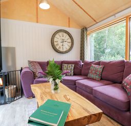A living room with a purple corner sofa wooden coffee table with a plant wood burning stove and large window at Lodge 22 in Corfe Castle
