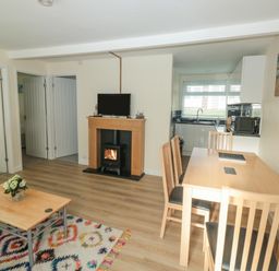 A room with wooden dining table and chairs a coffee table with flowers a fireplace with a stove and a television and a kitchen area at The Bolthole in Dartmouth