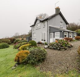 A two-story house with a grey exterior surrounded by a garden with shrubs and a gravel pathway at Sibrwd Y Mor in Harlech