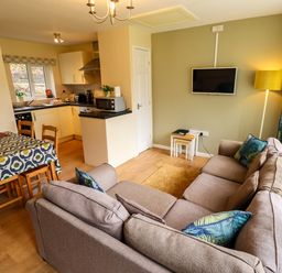 A living room with a brown sofa, a wall-mounted TV, a dining table with patterned tablecloth, and a kitchen area at 11 The Dell in Mundesley