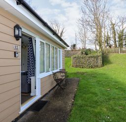 An exterior view of a beige house with an open door showing a glimpse of the inside and a small table with two chairs on a concrete patio next to a grass lawn at 12 The Dell in Mundesley