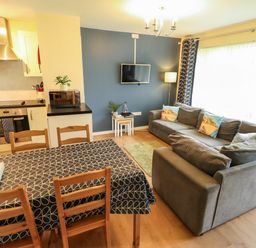 A living room with grey sofas a small green rug and a wall-mounted tv next to a dining table with four wooden chairs and a kitchen area at 12 The Dell in Mundesley