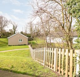A garden area with a wooden fence a small building and a larger white house surrounded by trees at 15 The Dell in Mundesley