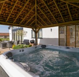 An outdoor hot tub under a wooden gazebo with patio furniture and a barbecue outside a house at Bilsham Meadows in Aust