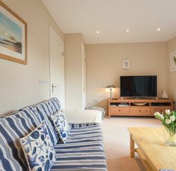 A living room with a striped sofa and patterned cushions a wooden TV cabinet a coffee table with flowers and framed pictures on the walls at Smugglers Cove 7 Beadnell