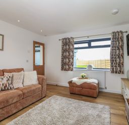 A living room with a brown sofa and patterned cushions a window with patterned curtains an ottoman with a tray and flowers a wooden door and a TV mounted on the wall at Refail Fach Near Cemaes Bay