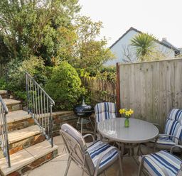 An outdoor patio with four cushioned chairs around a table with flowers and a barbecue grill next to stairs and plants at Bumbles in Padstow