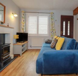 A living room with a blue sofa yellow pillows a wood stove a television on a stand and a window with patterned curtains at Bumbles in Padstow