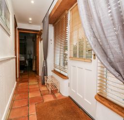 A hallway with terracotta floor tiles a white door with wooden blinds grey curtains a small wooden shoe rack and a framed picture on the wall at The Shippen in Gooseham near Kilkhampton