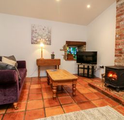 A living room with a purple sofa a wooden coffee table a small table with a lamp a television a wood burning stove and a stained glass window at The Shippen in Gooseham near Kilkhampton