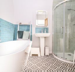 A bathroom with a white bathtub blue tiled walls a white sink with mirror a glass shower enclosure and patterned floor tiles at Sea Mouse Cottage Southwold