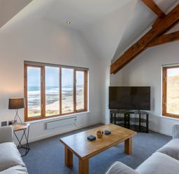 A living room with two sofas a wooden coffee table a television and windows showing a beach view at Sunset View in Widemouth Bay