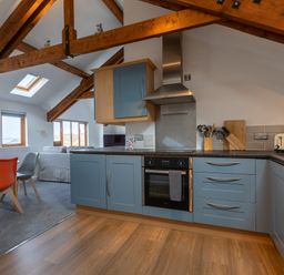 A kitchen with blue cabinets and wooden beams next to a dining area with chairs and a sofa at Sunset View in Widemouth Bay