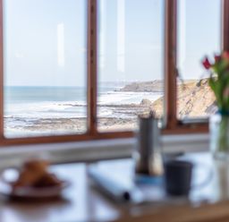 A coastal view from a window with table in foreground holding a cup flowers and plate at Sunset View in Widemouth Bay