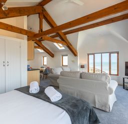 A room with a bed with towels a seating area a dining table wooden ceiling beams and large windows showing the sea at Sunset View in Widemouth Bay